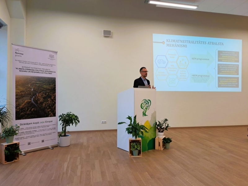 Lektors uzstājas konferenču telpā pie pults, fonā redzams prezentācijas slaids par klimata neitralitātes atbalsta mehānismu un informatīvs stends.