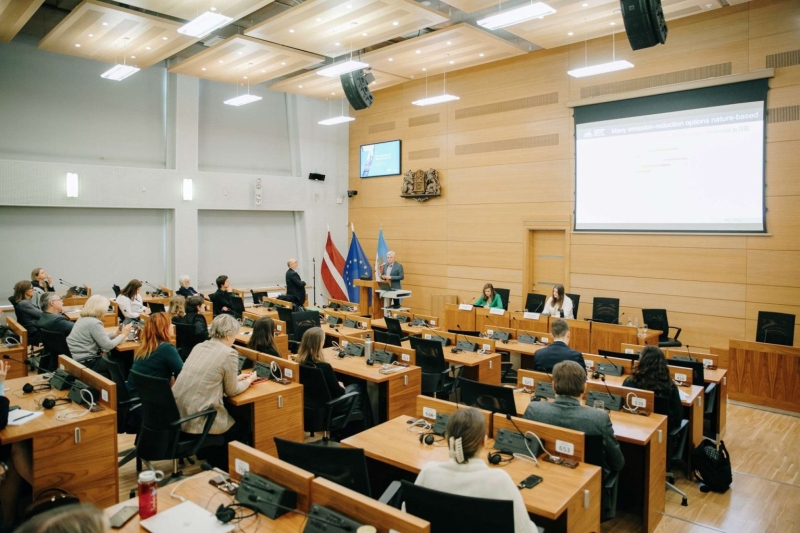 Zāle ar dalībniekiem, kas sēž pie galdiem un klausās prezentāciju pie ekrāna