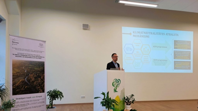 Lektors uzstājas konferenču telpā pie pults, fonā redzams prezentācijas slaids par klimata neitralitātes atbalsta mehānismu un informatīvs stends.