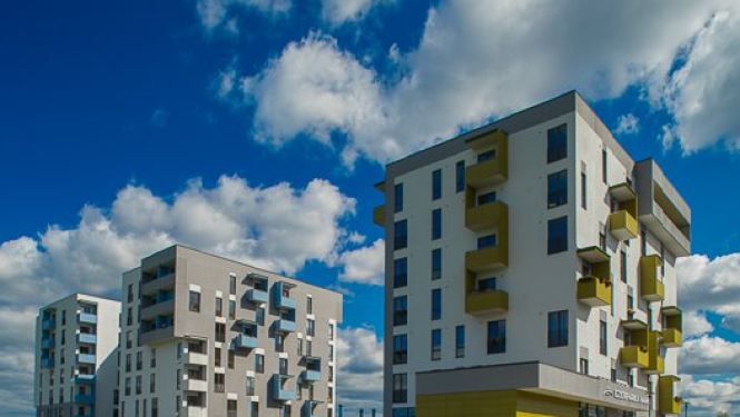 Mūsdienīgi daudzdzīvokļu dzīvojamie nami ar balkoniem zaļā apkārtnē, priekšplānā zāliens un gājēju celiņš, fonā zilas debesis ar mākoņiem.