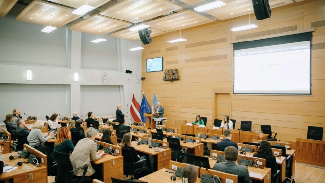 Zāle ar dalībniekiem, kas sēž pie galdiem un klausās prezentāciju pie ekrāna