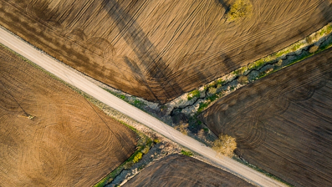Skats no augšas uz aramzemes laukiem, kurus pa diagonāli šķērso grants ceļš; gar ceļu starp laukiem vijas šaura ūdenstece ar veģetāciju.
