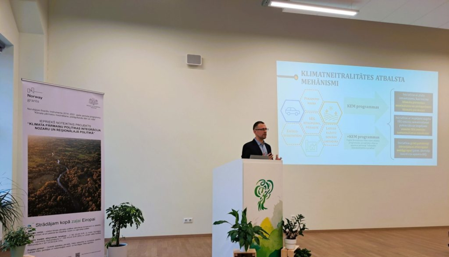 Lektors uzstājas konferenču telpā pie pults, fonā redzams prezentācijas slaids par klimata neitralitātes atbalsta mehānismu un informatīvs stends.