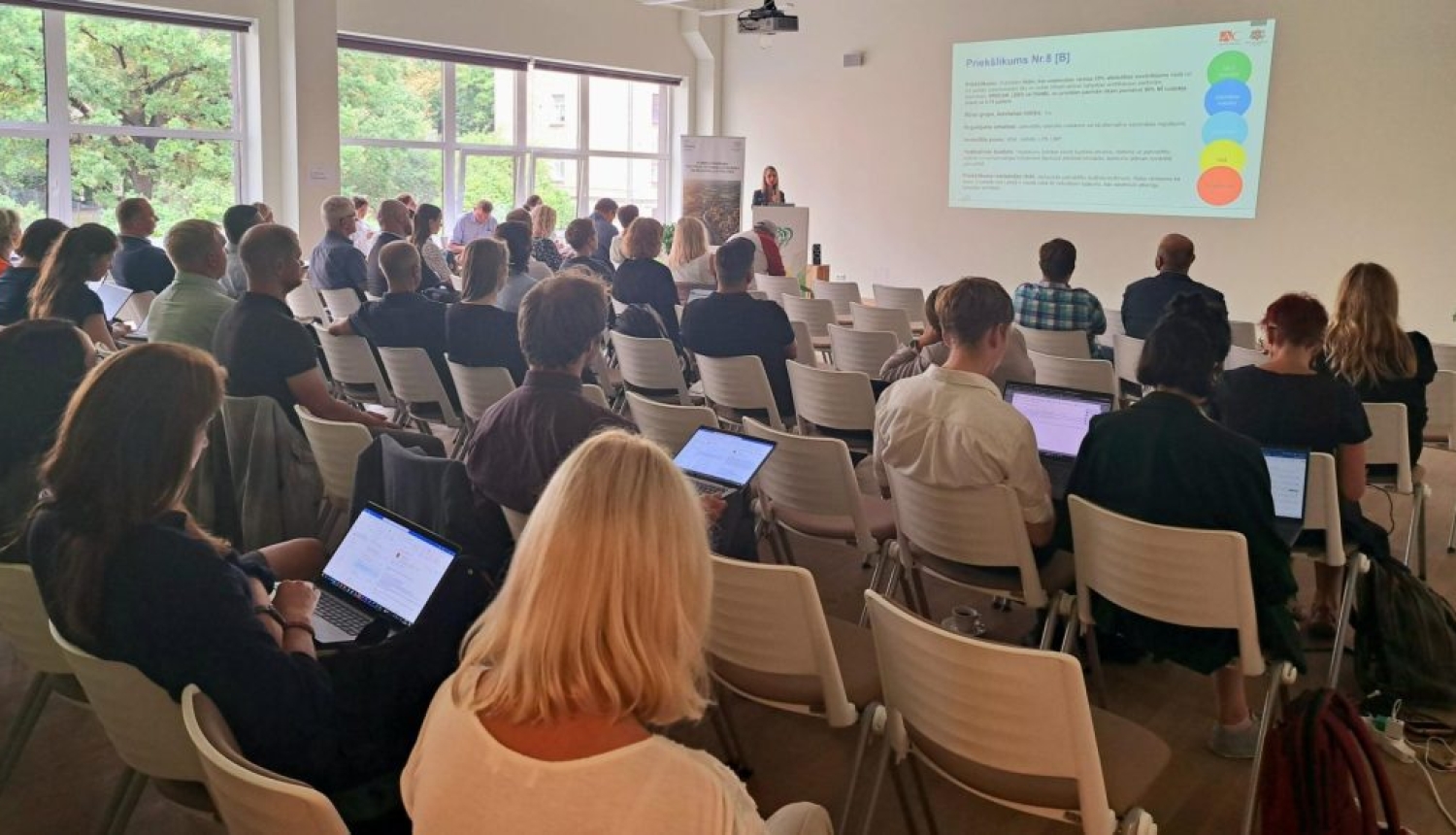 Prezentācija konferenču telpā ar klausītājiem un ekrānā redzamu slaidu.