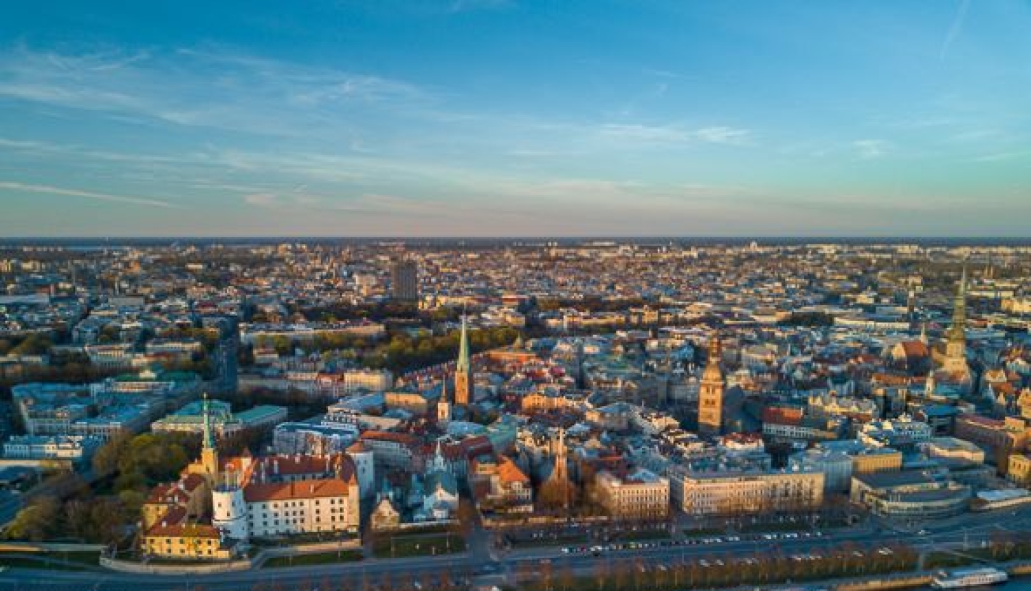 Skats no augšas uz pilsētas centru pie upes; redzama vēsturiskā apbūve, torņi, ielas un krastmala ar kuģiem saulainā laikā.